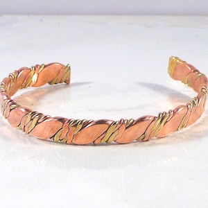May include: A copper bracelet with a twisted design. The bracelet features alternating sections of copper and gold-colored metal, creating a textured, open cuff design. The jewelry is displayed on a white surface.