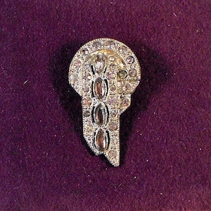 May include: A vintage silver-toned brooch with a geometric design. The brooch is encrusted with clear rhinestones, including three oval stones in the centre. The brooch has a rounded top and a stylized, asymmetrical shape.