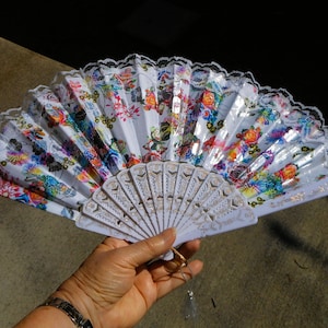 May include: A white hand fan with a floral design in various colors, including red, blue, and yellow. The fan has a white lace trim and is held open. The handle is white with gold accents.