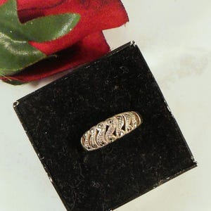 May include: A silver-toned ring with a decorative design featuring small, sparkling stones. The ring is set on a black velvet surface inside a small black box, with a red rose in the background.