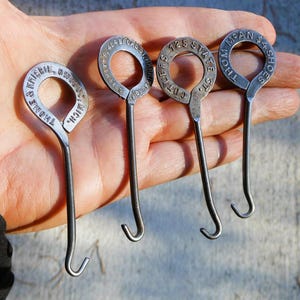 May include: Four vintage metal key rings with circular tops and curved hooks. Each ring has stamped text, including addresses and names. The rings are held in a hand, showcasing their aged, metallic appearance.