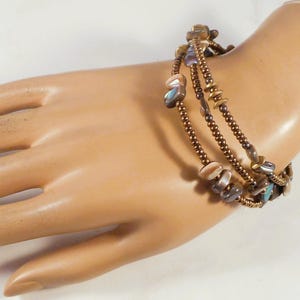 May include: A multi-strand bracelet with bronze-colored beads and decorative shell-shaped accents. The bracelet is wrapped around a wrist, showcasing its design and color variations. The bracelet's design includes small, round beads and larger, irregular-shaped shell pieces.