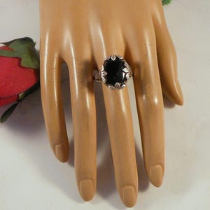 May include: A silver ring featuring a large, oval-shaped black stone set in a decorative metal frame with leaf-like engravings along the edges. The band is polished silver, and the setting holds the dark, smooth stone securely in place.