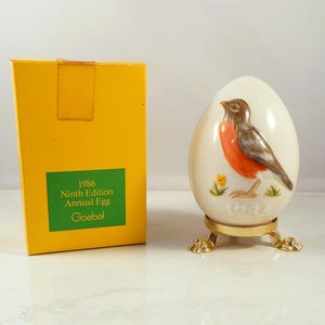 Puede incluir: Un huevo de cerámica decorativo con un diseño de pájaro pintado, colocado sobre un soporte dorado. El huevo es blanco con un pájaro marrón, naranja y gris. Se ve una caja amarilla con el texto verde "1986 Ninth Edition Annual Egg Goebel".