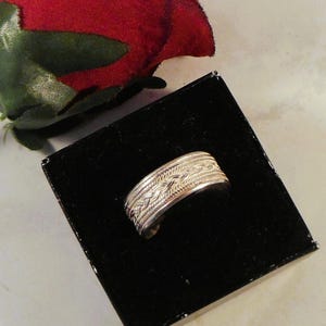 May include: A silver ring with a braided design, presented in a black box. The ring's intricate pattern is highlighted against the dark background. A red rose is partially visible in the background, adding a touch of color.