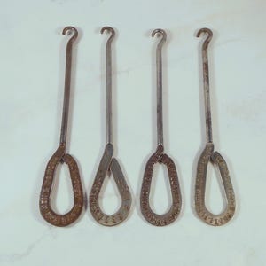 Könnte beinhalten: Vier antike Metall-Knopfhaken mit einem Haken oben und einer Schlaufe unten. Die Schlaufen haben Text an den Rändern. Die Knopfhaken sind silberfarben und haben ein Vintage-Aussehen.