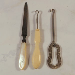 Peut inclure: Trois outils anciens : une lime à ongles en métal avec un manche en nacre, un crochet avec un manche crème et un crochet à bouton en métal avec le texte "E-Z CLAMP" et "QUALITY" sur la poignée.