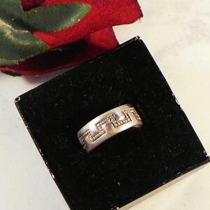 Puede incluir: Un anillo plateado con un patrón geométrico, exhibido en una caja negra. El anillo tiene una banda con un diseño repetitivo de formas conectadas. Se ve una tela roja en el fondo.