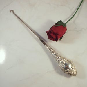 Può includere: Un uncinetto d'argento con un manico ornato e un'estremità a gancio ricurva. Una rosa rossa con uno stelo verde è accanto all'uncinetto. L'uncinetto è su una superficie bianca.