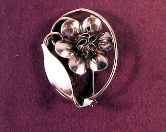 May include: A silver-toned metal brooch featuring a central flower with rounded petals and a detailed stamen, accompanied by a single smooth leaf. The entire floral arrangement is framed by a thin, oval-shaped metal wire loop.