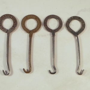 Könnte beinhalten: Vier vintage Knopfhaken aus Metall mit kreisförmigen Griffen. Jedes Werkzeug hat einen langen, dünnen Schaft, der in einem gebogenen Haken endet. Die kreisförmigen Griffe zeigen geprägten Text, darunter Thom McAn $4 Shoes auf dem ersten.