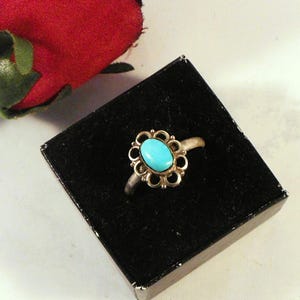 May include: A silver ring with a turquoise oval stone set in a gold-colored floral design. The ring is displayed on a black velvet surface, with a red rose in the background.