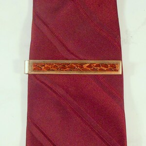 May include: A gold-toned metal tie clip featuring a rectangular inlay with a textured, reddish-brown snakeskin pattern. The tie clip is fastened horizontally across a solid, deep burgundy necktie that has subtle diagonal woven stripes.