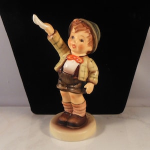 May include: A ceramic figurine of a young boy in a plaid jacket, shorts, and a cap, with a red bow tie. The boy is standing on a round base, with one arm raised. The figurine has a vintage aesthetic.