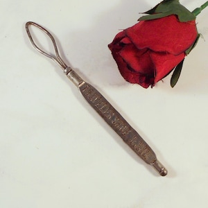 Könnte beinhalten: Ein vintage Knopfhaken aus Metall mit einer geschlungenen Drahtspitze und einem flachen Griff. Der Griff ist mit dem Text TRAVELER SHOE J. WITT MAKER geprägt. Das Werkzeug hat eine angelaufene silberfarbene Oberfläche und liegt diagonal neben einer roten Stoffrose.