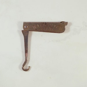 Könnte beinhalten: Ein Vintage-Schuhhaken aus Metall mit dem Text "WALK-OVER SHOE" auf der Oberseite. Der Haken hat eine bräunlich-bronze Farbe und einen gebogenen Haken am Ende. Der Haken ist offen und einsatzbereit.