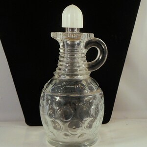 May include: Clear glass cruet with a white stopper and a handle. The cruet has a textured, bubble-like pattern on the bottom half and a ribbed design on the neck. The handle is a loop shape. The cruet is designed for holding liquids.