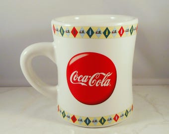 May include: White ceramic Coca-Cola mug with a large red circle featuring the Coca-Cola logo in white script. The mug has a decorative band of colorful diamond shapes around the top and bottom.