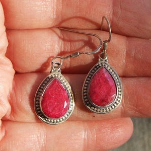 May include: A pair of silver-toned drop earrings with a teardrop-shaped, faceted, red gemstone center. The earrings have a beaded border and a simple hook closure. The gemstones are a vibrant shade of red.