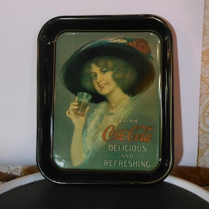 May include: Vintage Coca-Cola serving tray with a black frame. The tray features an illustration of a woman in a hat holding a glass of Coca-Cola. The text on the tray reads "DRINK Coca-Cola DELICIOUS AND REFRESHING."