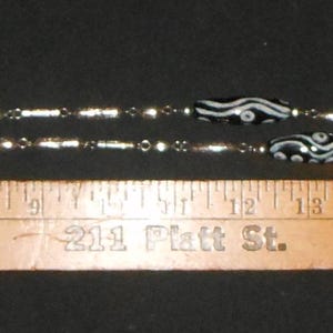 Peut inclure: Un collier argent&eacute; orn&eacute; de perles noires et blanches &agrave; motifs. Le collier est pos&eacute; &agrave; c&ocirc;t&eacute; d'une r&egrave;gle en bois avec le texte "GROSS AUTOMOTIVE" et un num&eacute;ro de t&eacute;l&eacute;phone. Le collier mesure environ 46 cm de long.