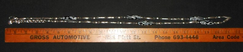 Peut inclure: Un collier argent&eacute; orn&eacute; de perles noires et blanches &agrave; motifs. Le collier est pos&eacute; &agrave; c&ocirc;t&eacute; d'une r&egrave;gle en bois avec le texte "GROSS AUTOMOTIVE" et un num&eacute;ro de t&eacute;l&eacute;phone. Le collier mesure environ 46 cm de long.