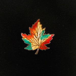 May include: A colorful maple leaf pin with a silver-tone border. The leaf is painted with shades of red, orange, yellow, and green. The pin is set against a black background.