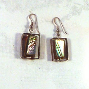 May include: A pair of silver earrings with rectangular iridescent abalone shell inserts. The shell displays a mix of colors, including green, blue, and gold. The earrings have a simple, modern design with a hook closure.