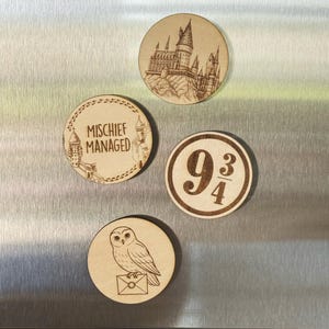 May include: Four round wooden magnets with etched designs. One features Hogwarts castle, another has the text "Mischief Managed", a third displays "9 3/4", and the last shows an owl with an envelope. The magnets are displayed on a stainless steel surface.