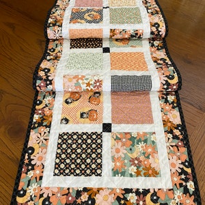 May include: A black and white table runner with a floral pattern and a patchwork design. The runner is made up of different squares with various patterns, including black and white polka dots, black and white gingham, and a floral pattern. The runner has a black border with a white trim.