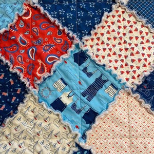 May include: Close-up of a patchwork quilt with various patriotic and summer-themed patterns. The quilt features red paisley, watermelon slices, sailboats, and swimwear designs. The edges are finished with a ruffled trim.