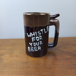 May include: A brown ceramic beer mug with a handle shaped like a deer's head. The mug has white lettering that reads "Whistle For Your Beer".