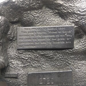 May include: A silver-toned metal belt buckle with a raised eagle design. The buckle has a plaque with the text "1989 PROUD TO SERVE 1990" and another plaque with the text "No. 2720 of 5,000 Limited Edition".