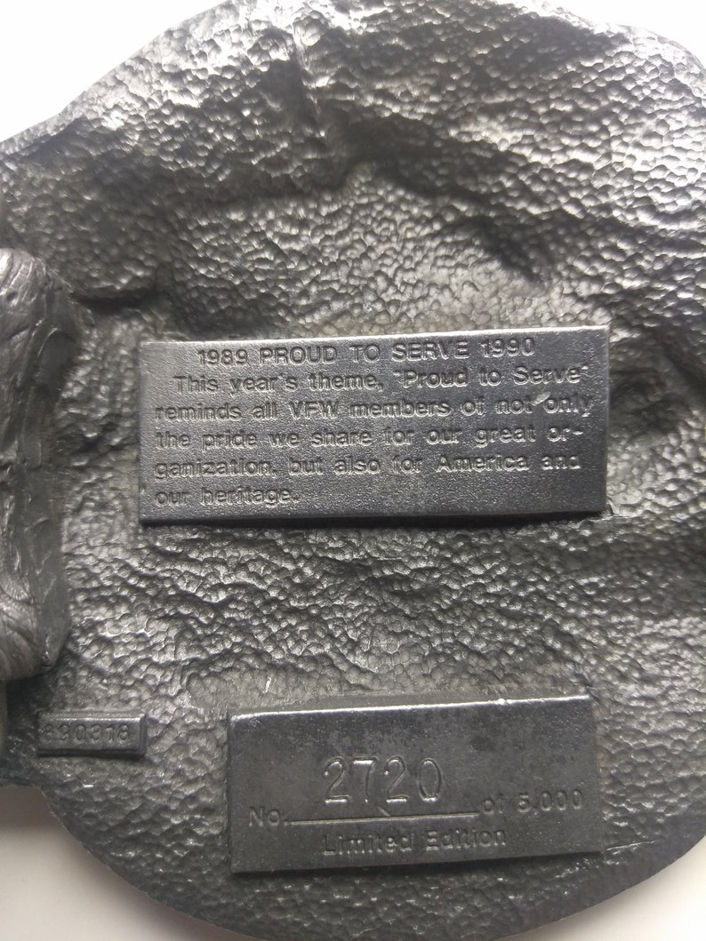 May include: A silver-toned metal belt buckle with a raised eagle design. The buckle has a plaque with the text "1989 PROUD TO SERVE 1990" and another plaque with the text "No. 2720 of 5,000 Limited Edition".