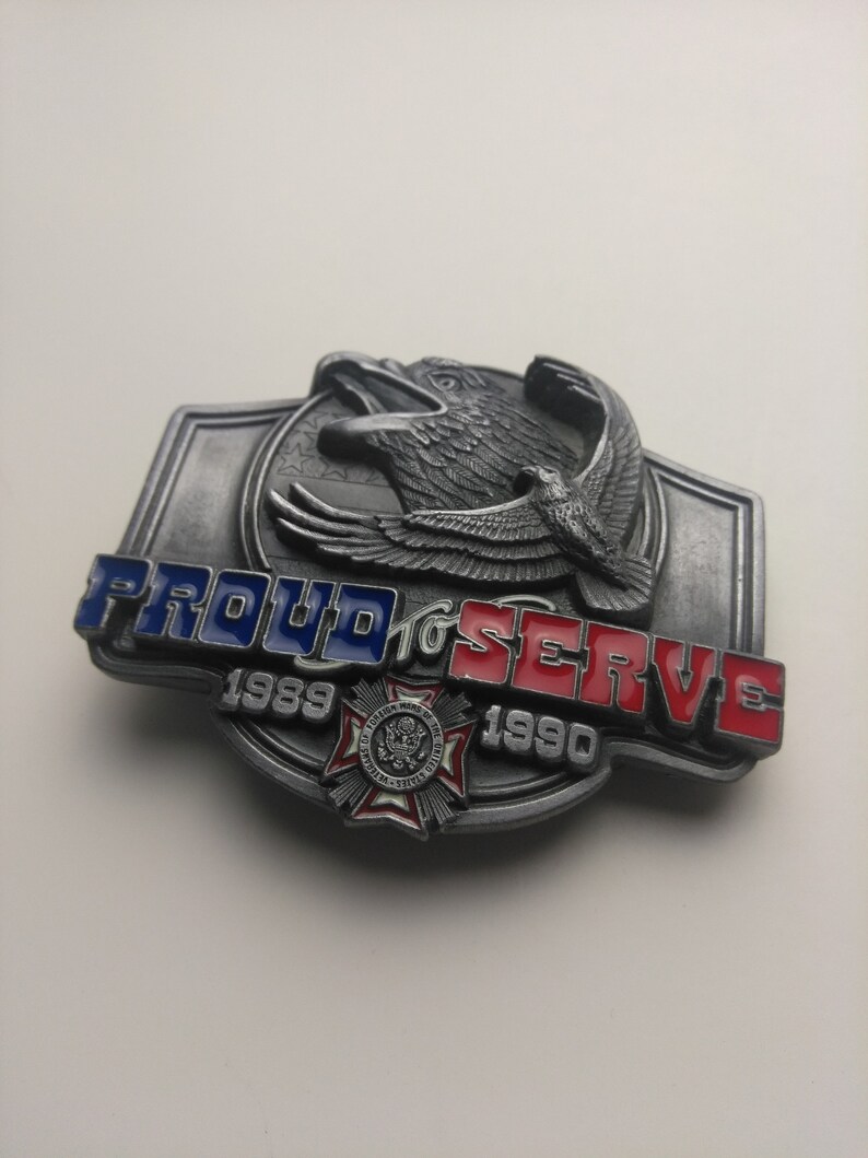 May include: A silver belt buckle with a red, white, and blue eagle design. The buckle says "Proud to Serve 1989-1990".
