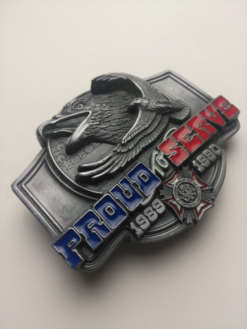 May include: A silver belt buckle with a bald eagle design and the words "Proud to Serve 1989-1990". The buckle also features a red, white, and blue American flag design and a small red, white, and blue emblem with the words "Veterans of the United States of America".