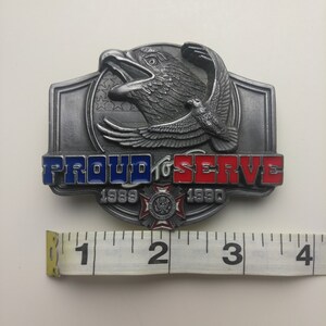 May include: A silver belt buckle with a raised eagle design and the words "PROUD TO SERVE" in red, white, and blue enamel. The buckle also features the years "1989" and "1990" and a small emblem.