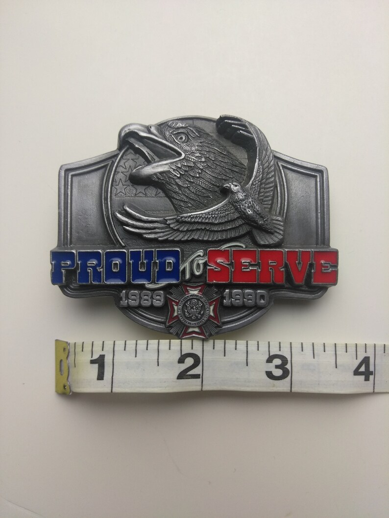 May include: A silver belt buckle with a raised eagle design and the words "PROUD TO SERVE" in red, white, and blue enamel. The buckle also features the years "1989" and "1990" and a small emblem.