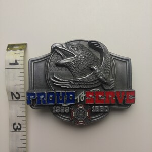 May include: A silver belt buckle with a bald eagle design. The buckle has the words "Proud to Serve" and the years "1989" and "1990" on it. The eagle is flying with its wings spread and has a red, white, and blue American flag in the background.