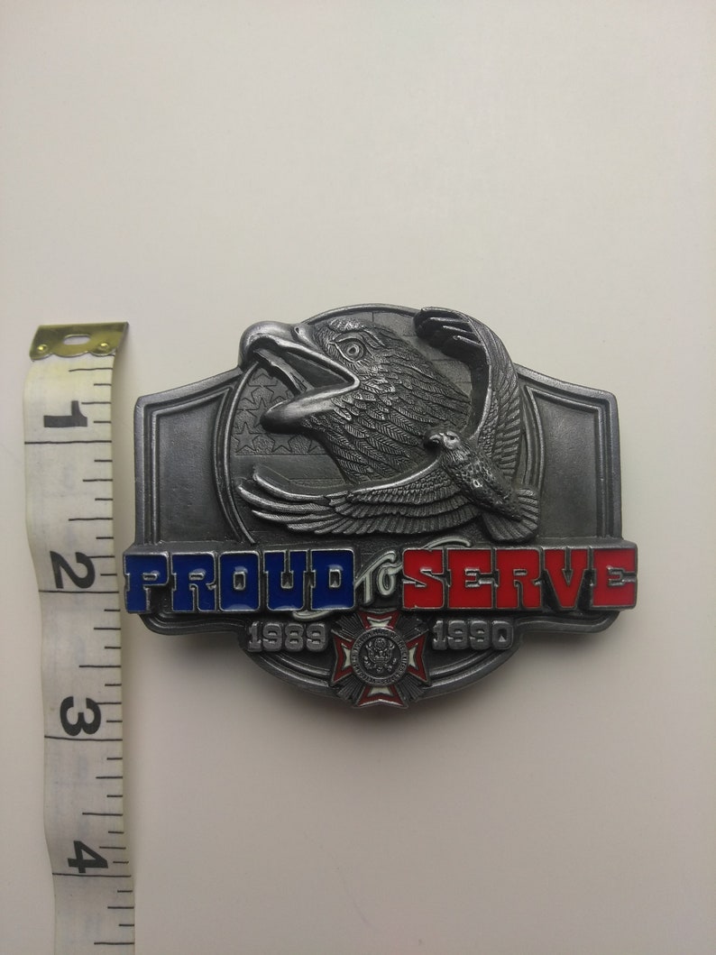 May include: A silver belt buckle with a bald eagle design. The buckle has the words "Proud to Serve" and the years "1989" and "1990" on it. The eagle is flying with its wings spread and has a red, white, and blue American flag in the background.
