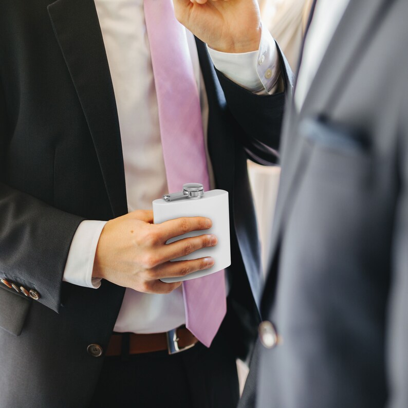 May include: A man in a black suit holds a white flask with a silver cap. He is wearing a pink tie and a white shirt.
