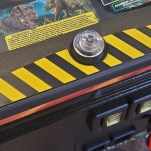 May include: A black and yellow striped warning sign with a clear plastic dome on top. The sign is attached to a black surface.