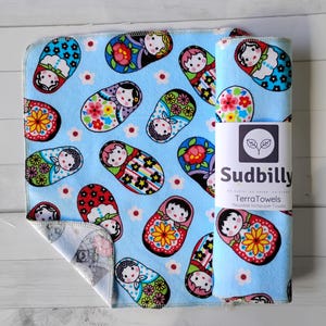 May include: Blue reusable TerraTowels with a repeating pattern of colorful Matryoshka dolls and small white flowers. One towel is rolled up with a label that reads "Sudbilly TerraTowels Reusable NoPaper Towels."