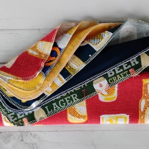 May include: A stack of beer-themed fabric napkins. The napkins feature various patterns, including beer bottles, beer mugs, and the words "Lager" and "Beer Craft." The colors include red, yellow, navy blue, and green.