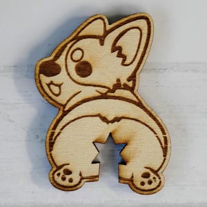 May include: Wooden corgi butt-shaped cutout. The cutout features a detailed outline of a corgi's rear, with a star-shaped cutout in the center. The wood has a light brown color with darker brown detailing, showcasing the corgi's features.