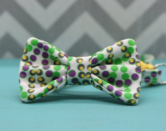 May include: A fabric bow tie with a white background and a pattern of green, purple, and yellow polka dots. The bow tie is centered on a light blue surface, with a gray and white chevron pattern in the background.