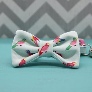 May include: A light blue bow tie featuring a repeating pattern of colorful birds. The birds are depicted in shades of pink, green, yellow, and blue. The bow tie is made of fabric and has a classic bow shape.