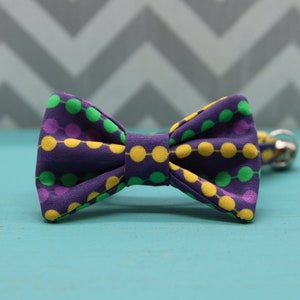 May include: A purple bow tie with a pattern of yellow, green, and purple circles. The bow tie is made of fabric and has a classic bow shape. The background is a light blue surface with a gray and white chevron pattern.