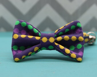 May include: A purple bow tie with a pattern of yellow, green, and purple circles. The bow tie is made of fabric and has a classic bow shape. The background is a light blue surface with a gray and white chevron pattern.