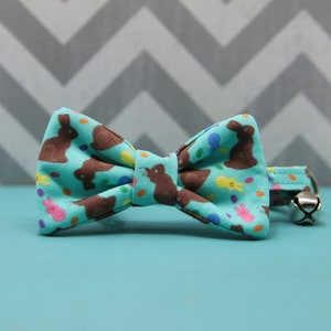 May include: A turquoise bow tie with a repeating pattern of brown Easter bunnies, colorful eggs, and chicks. The bow tie is attached to a matching collar with a silver bell, perfect for a pet.